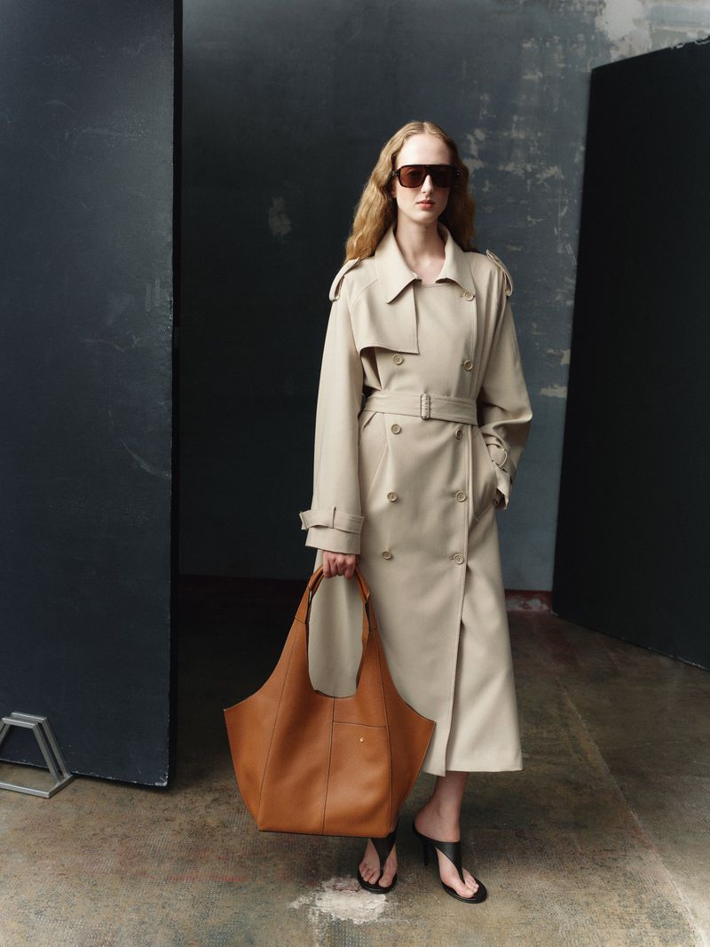 A stylish adult woman wears a beige trench coat and sunglasses, carrying a large brown leather tote bag in a studio setting.