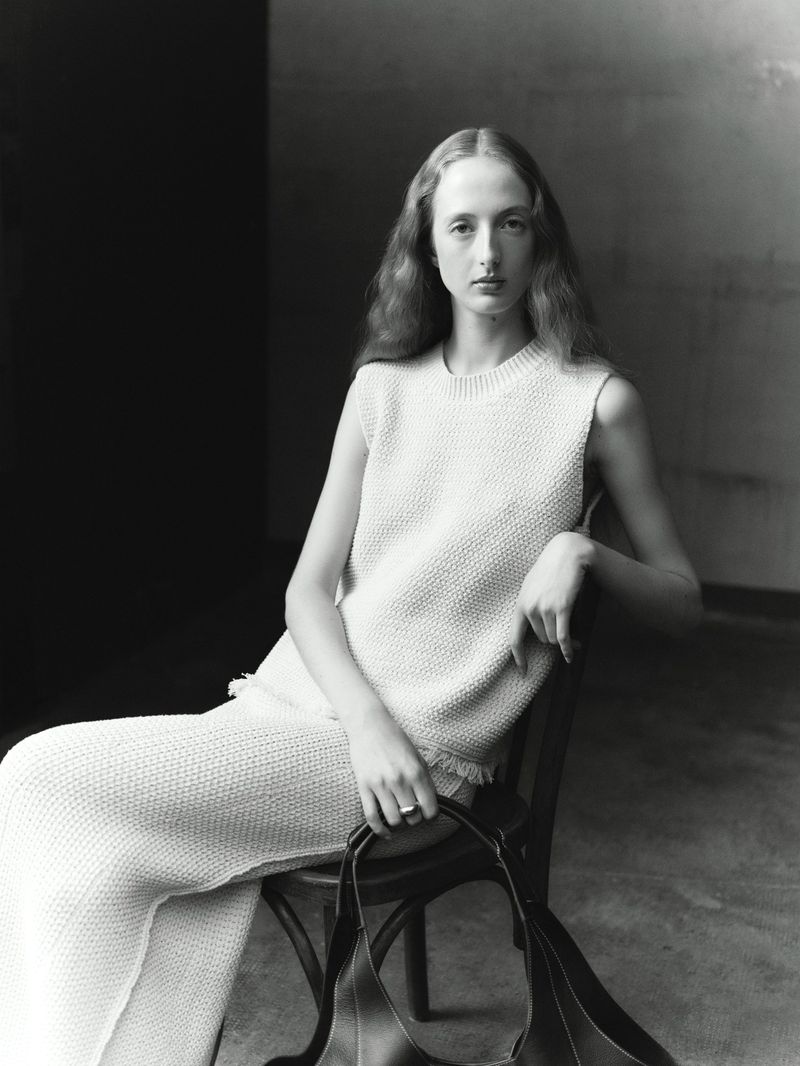 Black and white portrait of an adult woman sitting on a chair, wearing a knit sweater and pants, and holding a bag.
