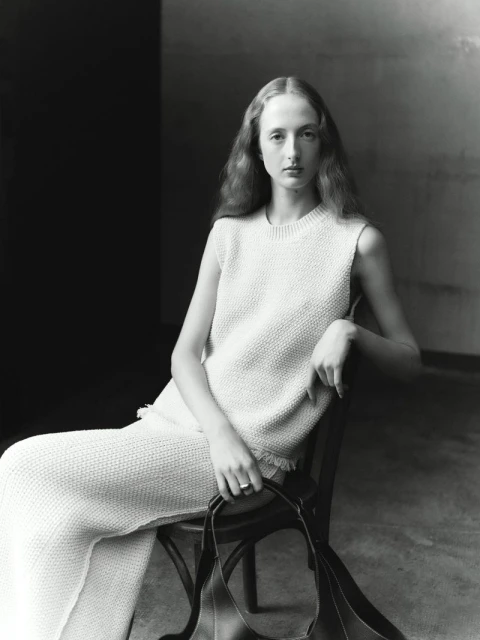 Black and white portrait of an adult woman sitting on a chair, wearing a knit sweater and pants, and holding a bag.