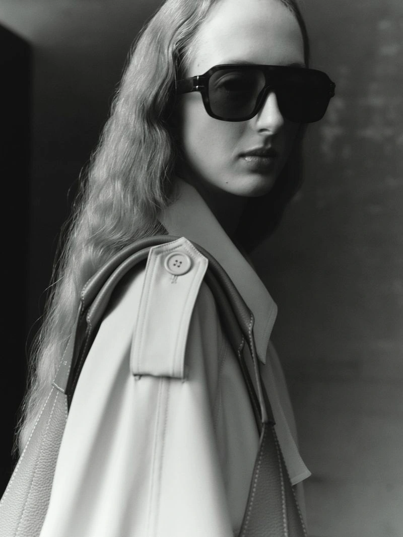 Model wearing dark oversized sunglasses and white trench coat with leather bag strap for RSVP Paris