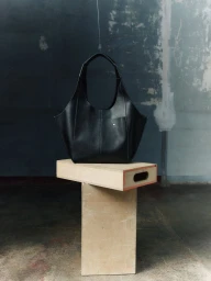 Sculptural black leather tote bag displayed on stacked wooden apple boxes against a textured distressed wall.