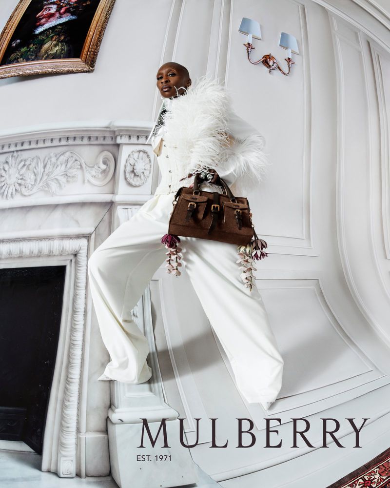 Fashion shot of a Black woman in white pantsuit and feathered boa holding a brown Mulberry bag against a painted fireplace backdrop.