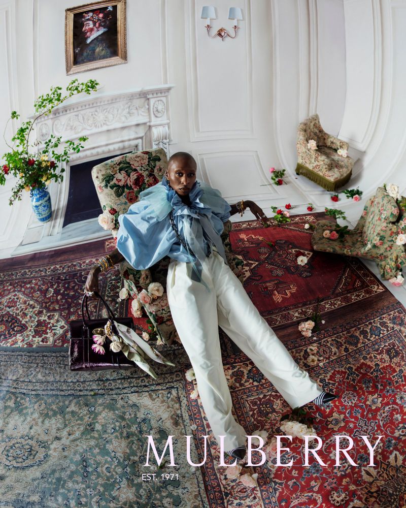 An adult female poses in a light blue top and white pants amidst scattered pink flowers and antique rugs inside an ornate room.