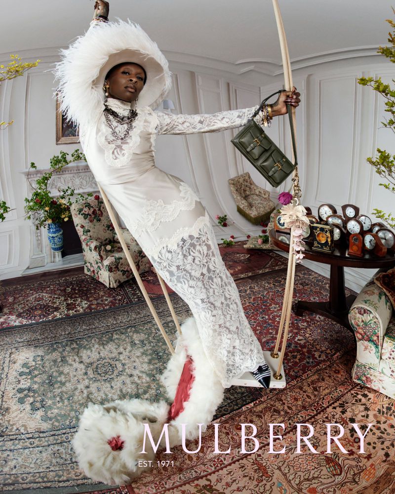 An adult woman in a lacy white dress and feathered hat poses on a swing indoors, holding a green purse, surrounded by antique furniture.