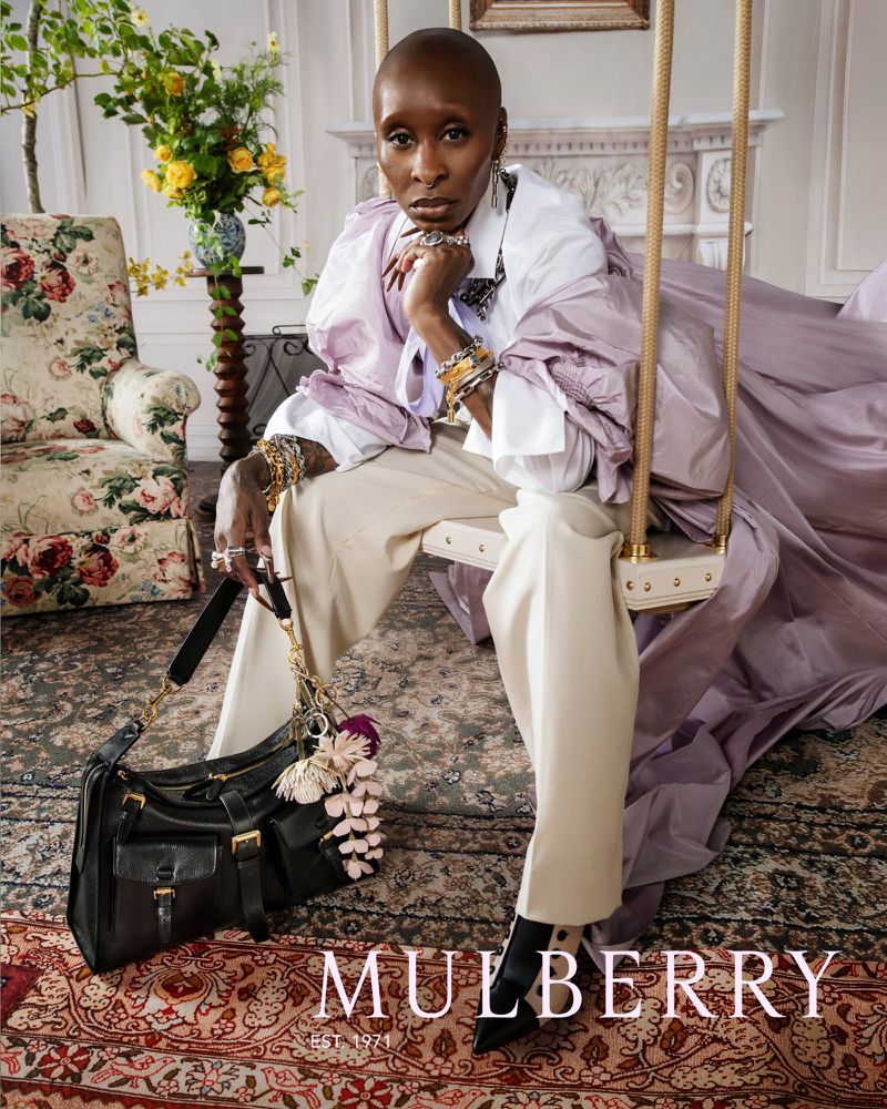 Woman in designer outfit sits on swing wearing jewelry, white button-down shirt, and tan slacks, with a Mulberry handbag and floral chair.