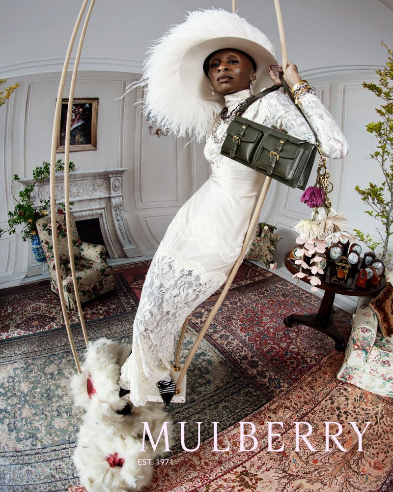 A woman wearing a white lace dress, a large feathered hat, and holding a Mulberry bag, posing in an ornate indoor setting on a swing.