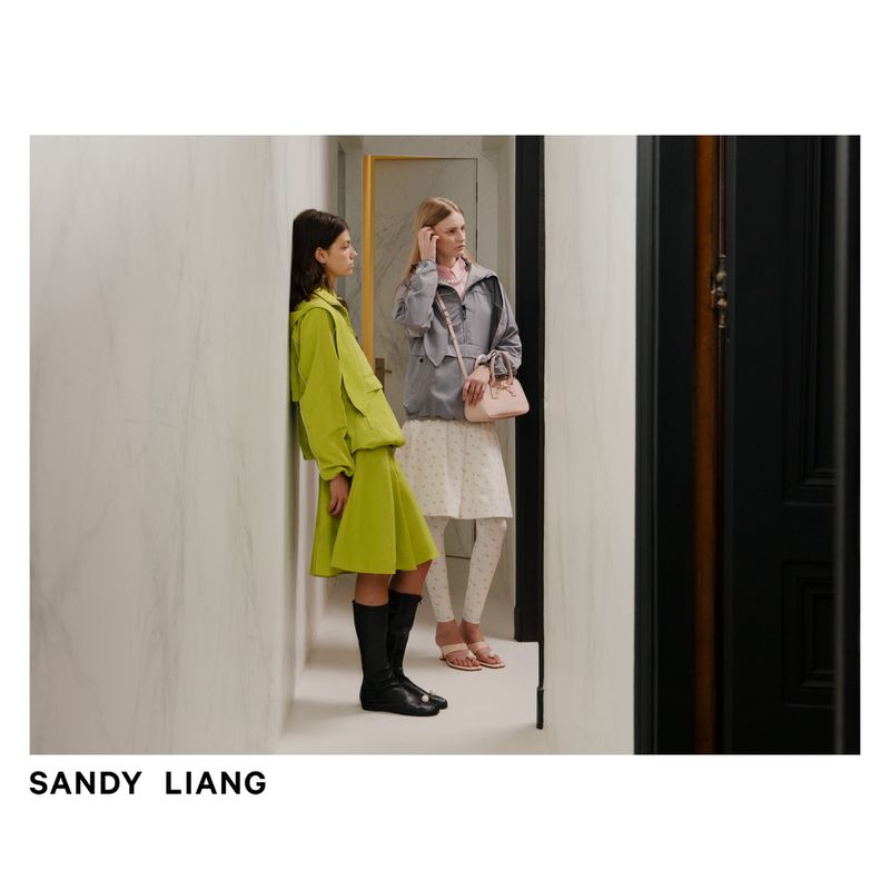 Two women in fashionable outerwear and dresses pose in a hallway; one in lime green, the other in grey, in a contemporary setting.