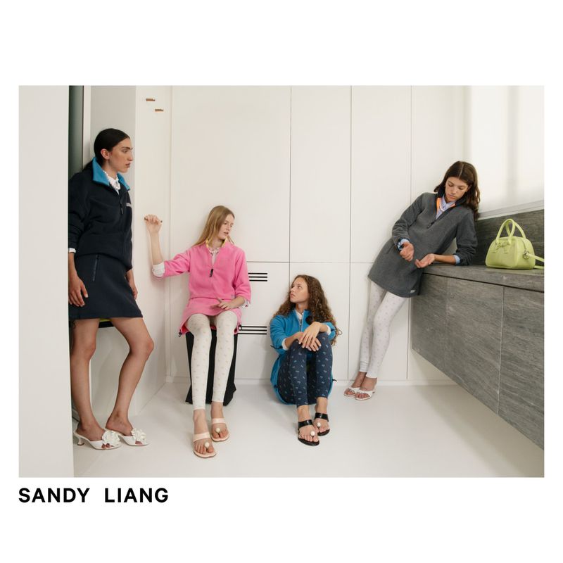 Four fashion models in a minimalist interior wear Sandy Liang apparel: colorful tops, leggings, sandals, and a green purse, capturing a modern style.
