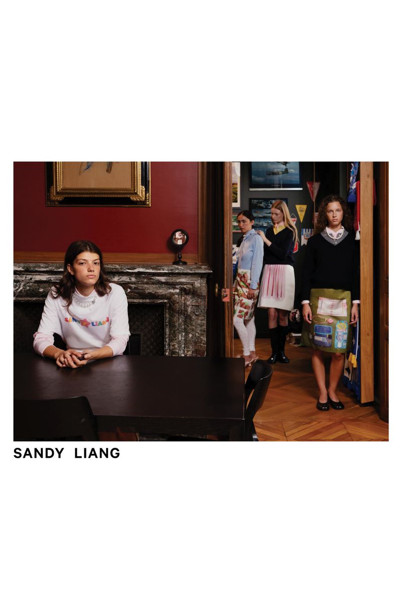 Fashion editorial shot of a woman seated at a table, with three women standing in a doorway in the background of an indoor space.