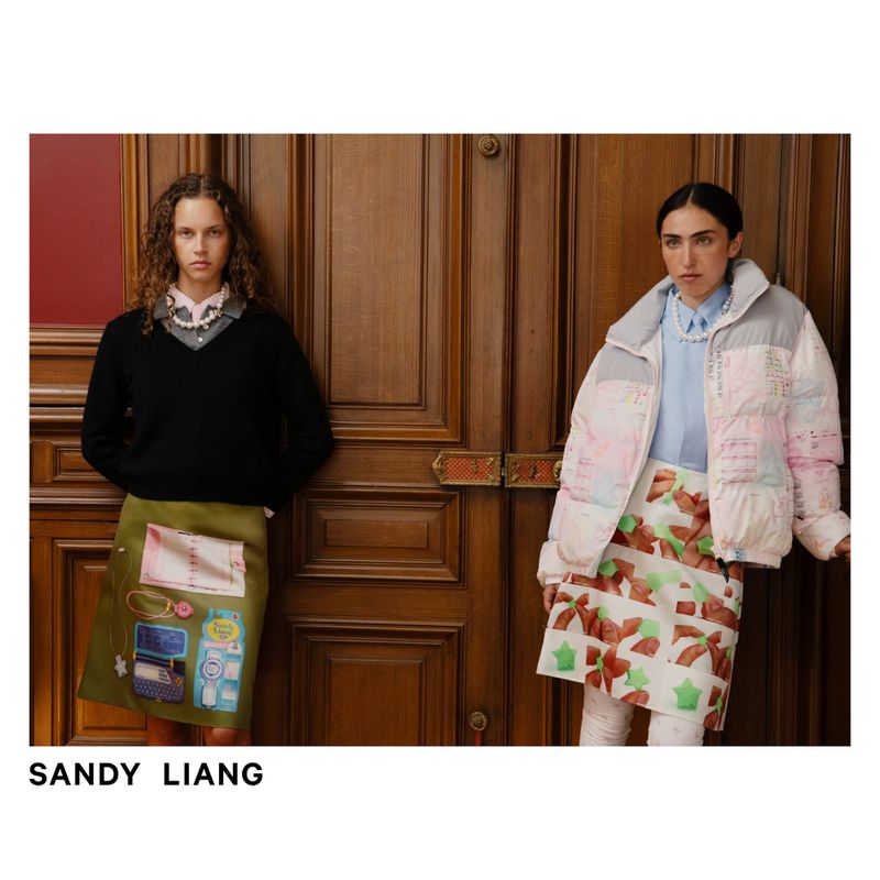 Two adult females pose in front of a wooden door in a fashion editorial. One wears a black sweater and the other a puffer jacket.