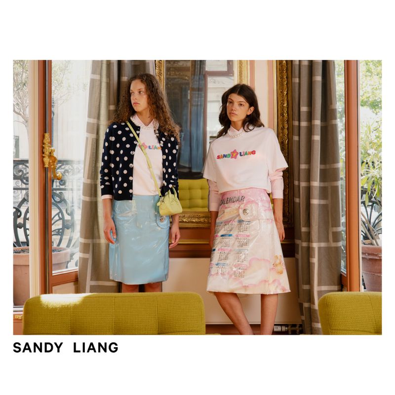 Two women model Sandy Liang skirts and T-shirts in front of a window with neutral curtains and architectural details, indoor shot.