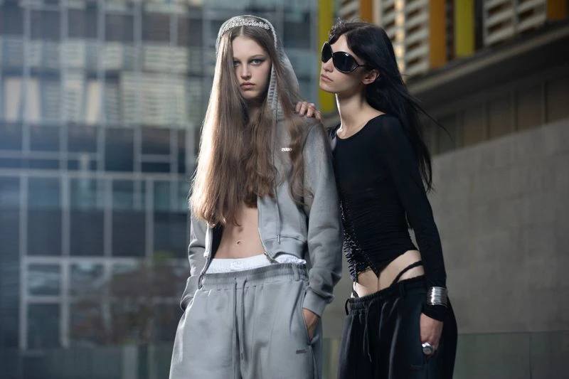 Two women pose outdoors in fashionable sportswear against a modern building backdrop, showcasing contemporary street style and athletic wear.