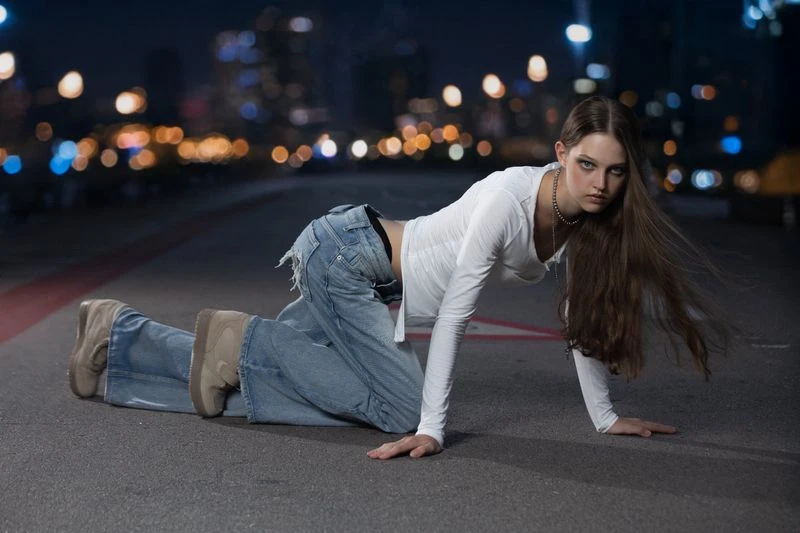 Young woman poses on all fours in ripped jeans and a long-sleeved white top, set against an urban cityscape at dusk with blurred lights.