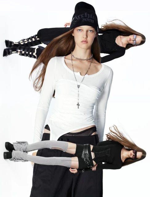 Fashion editorial shot featuring a fair-skinned woman in a white long-sleeve top, set against a clean white backdrop with two reflected figures.