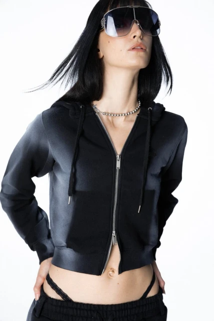 Brunette woman in sunglasses and hoodie in a white studio. Close-up portrait with cool lighting, showcasing fashion and modern style.
