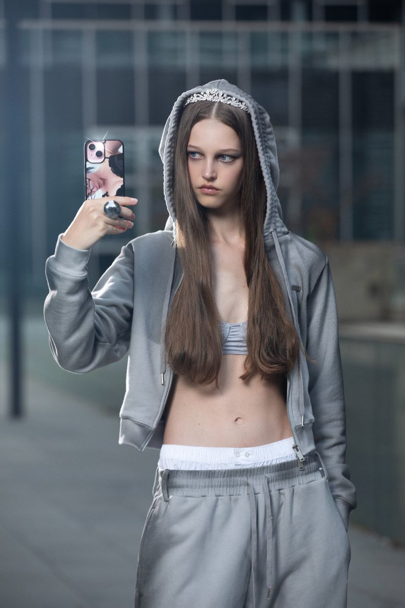 Teen girl in a gray tracksuit and tiara taking a selfie with her cell phone outdoors in front of a modern building. Fashion style.