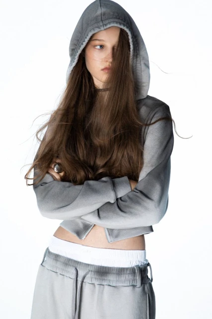 Young woman in gray hoodie and sweatpants posing with crossed arms against a white background in a studio. Fashionable portrait.