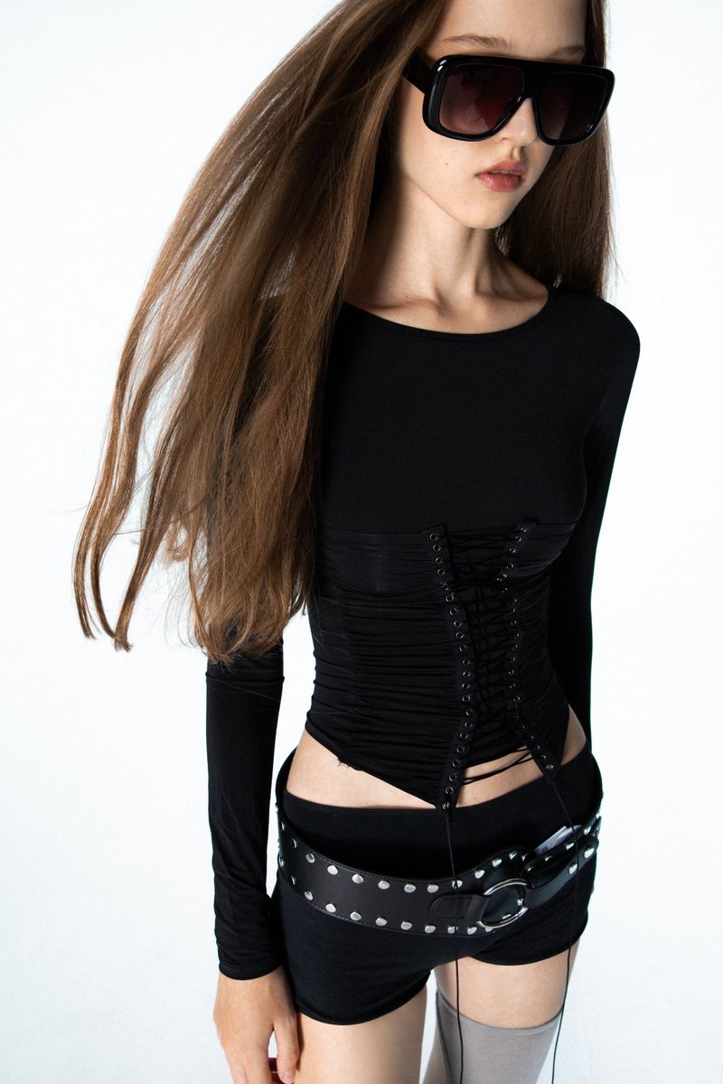 Model wearing oversized dark sunglasses, a black lace-up corset top, and a wide studded belt against a white studio background.