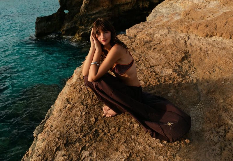 A woman in a brown bikini top and long skirt sits on a rocky ledge by the turquoise sea, posing with hands near her face, looking at the camera.