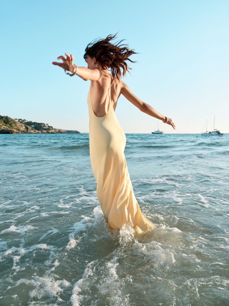 Mia Regan wades into the ocean wearing a long pale yellow backless slip dress with arms outstretched.
