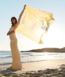 Model Mia Regan on a sandy beach holding up a large yellow patterned scarf against the sunlight.