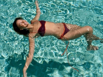 Model Mia Regan floating in a clear turquoise pool wearing a burgundy two-piece bikini with orange trim.