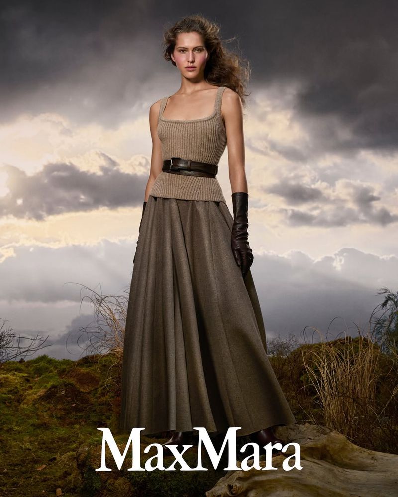 Fashion shot of a woman wearing a beige top, long skirt, and gloves, posing outdoors against a cloudy sky. Max Mara logo at the bottom.