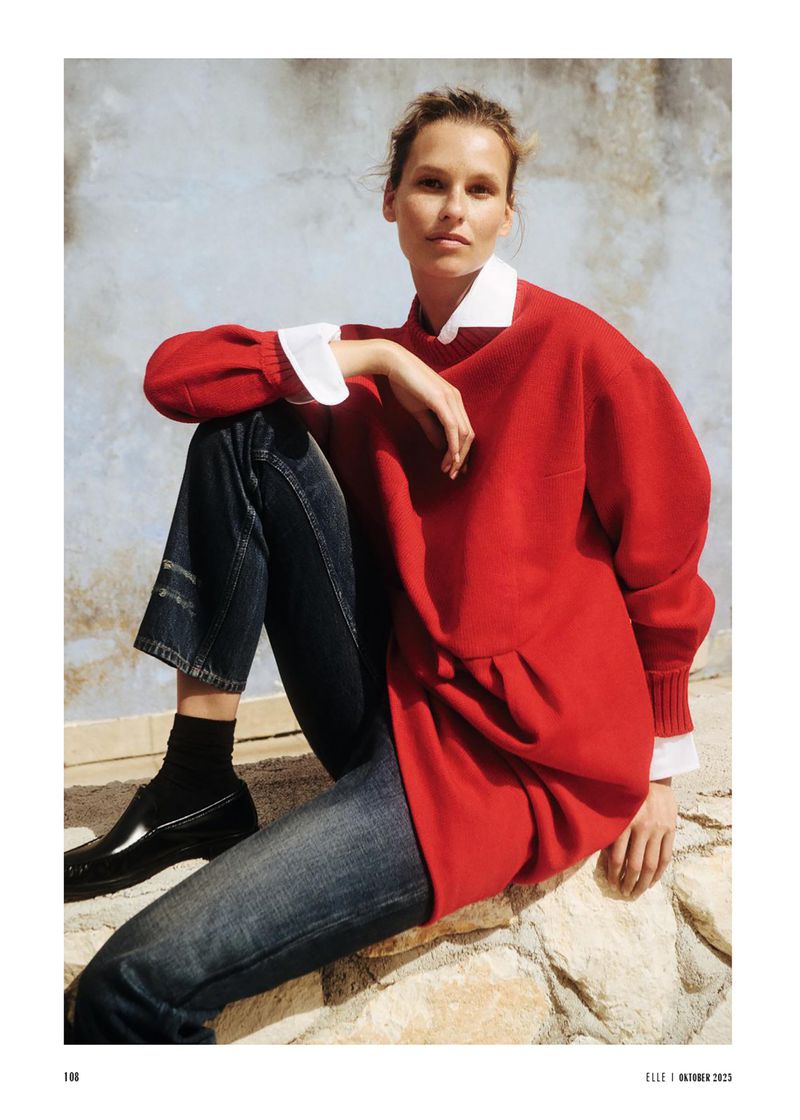 Fashion editorial shot of a woman in a red sweatshirt, dark denim pants, and shiny black shoes sitting outside on a stone surface.