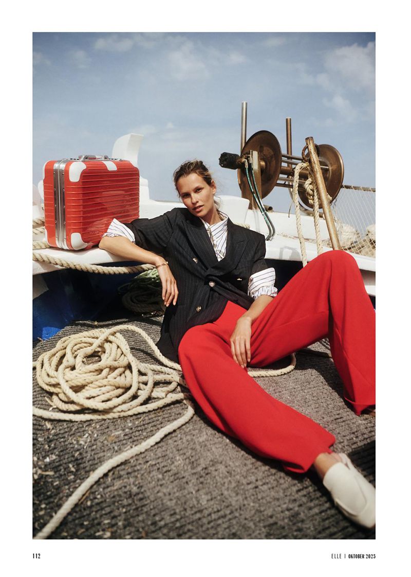 Fashion shot of a woman in a blazer and red pants sitting on a boat next to a red suitcase with a rope, nautical theme, cloudy skies.
