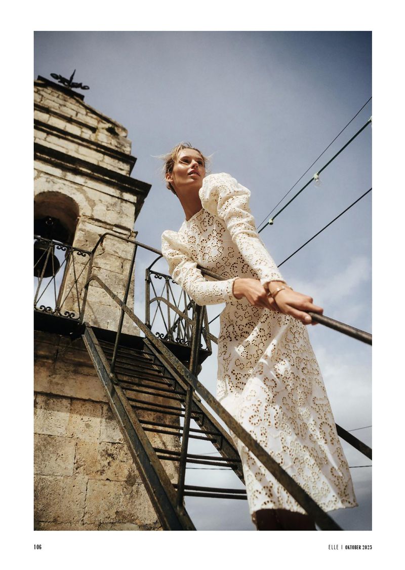 Fashion shot of a woman in a long-sleeved dress leaning against a railing by a stone building under a bright, sunny sky.