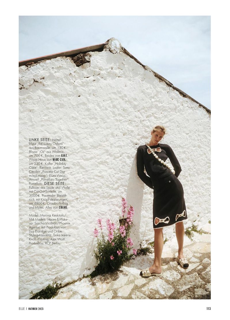Fashion editorial shot of a female model in a black skirt and sweater with bow details against a white stone wall backdrop.