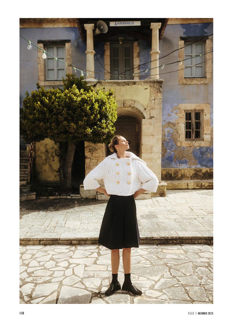 A woman in a white jacket and black skirt stands on a stone surface in front of a weathered building with columns and archways.