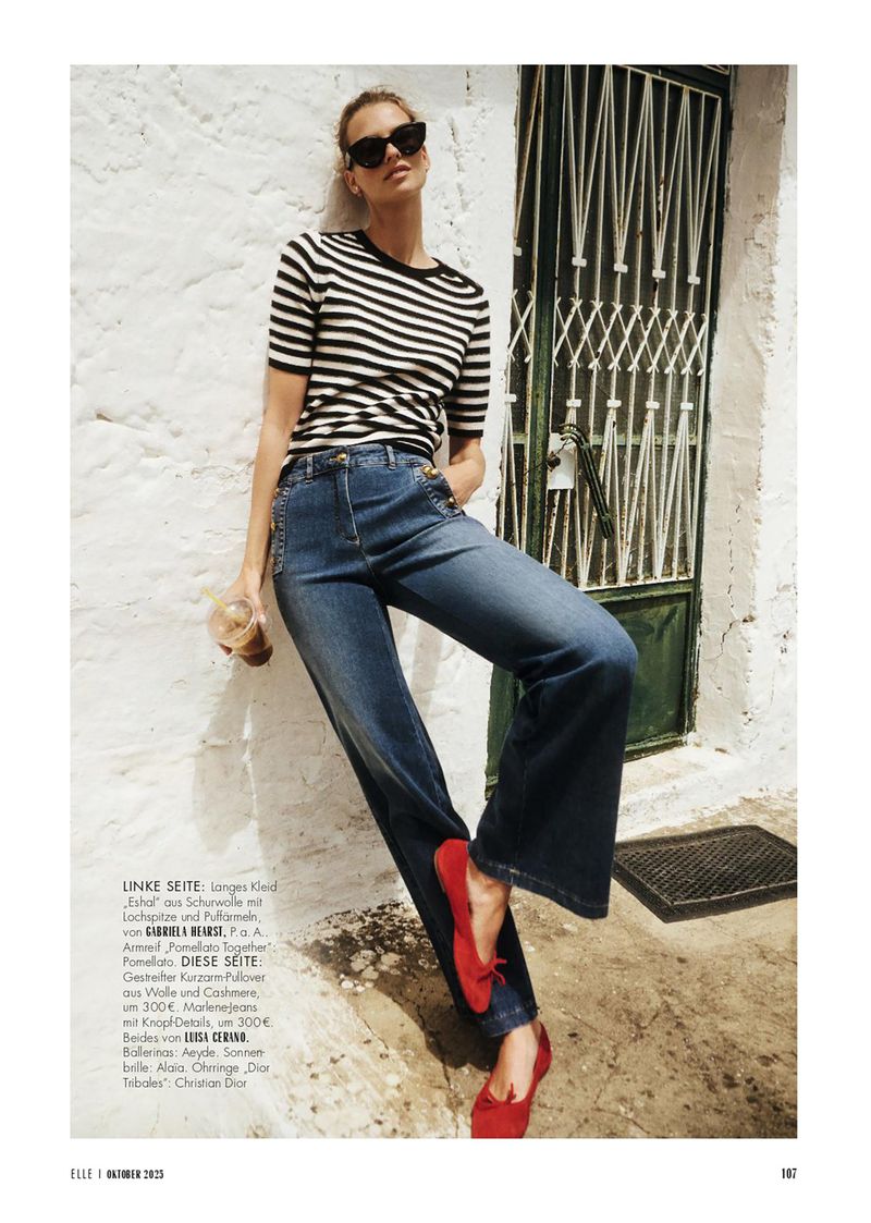 Fashion editorial shot: Woman in striped top, jeans, and red flats leans against a white wall with ornate doorway; holding iced coffee.
