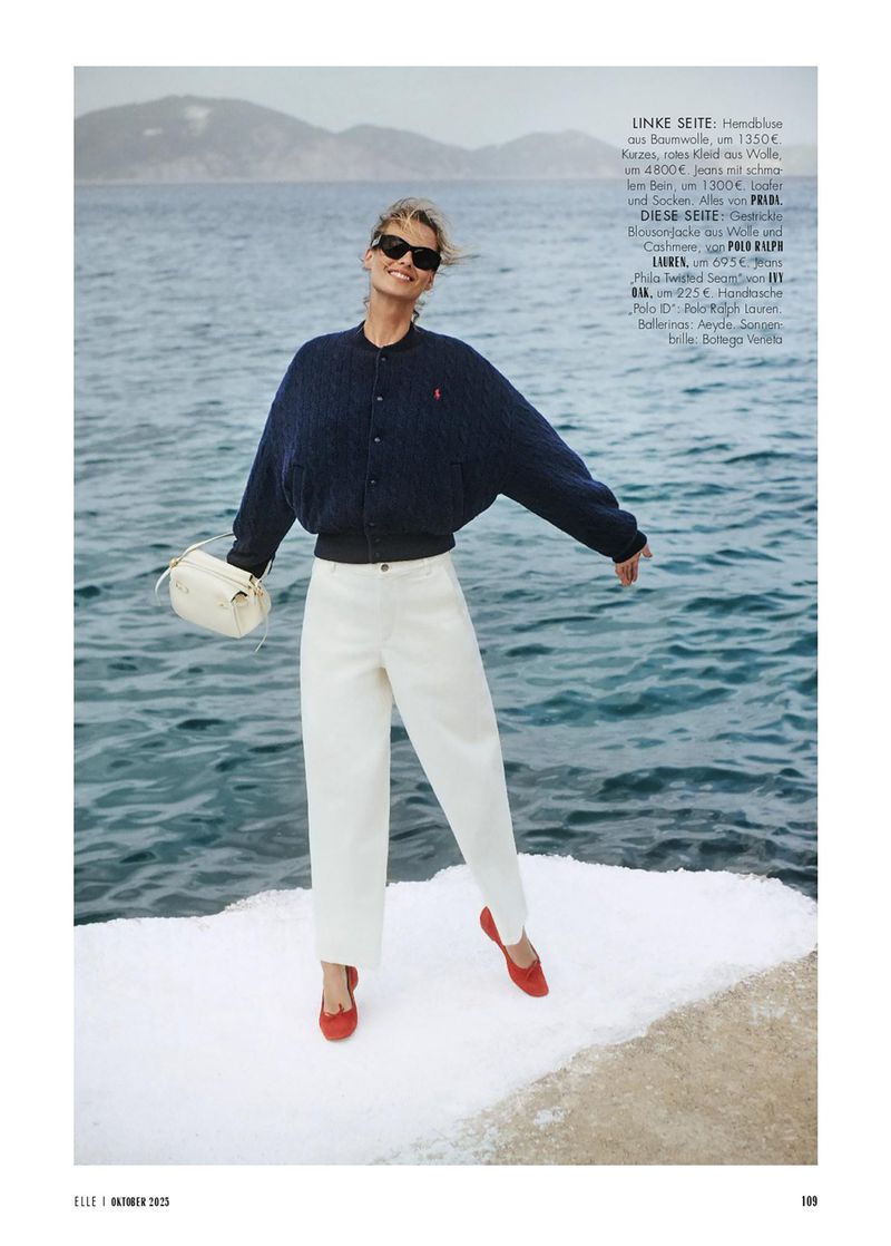 Blonde woman in a blue bomber jacket, white pants, and red flats poses in front of the ocean holding a white purse.