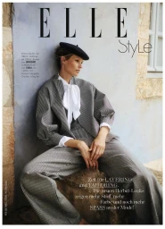 Model in grey striped suit and black beret seated against rustic stone wall for Elle Germany magazine editorial shoot