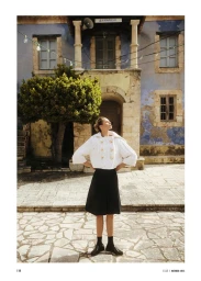 Model standing outdoors in a white knitted buttoned jacket and black skirt before a rustic stone building.