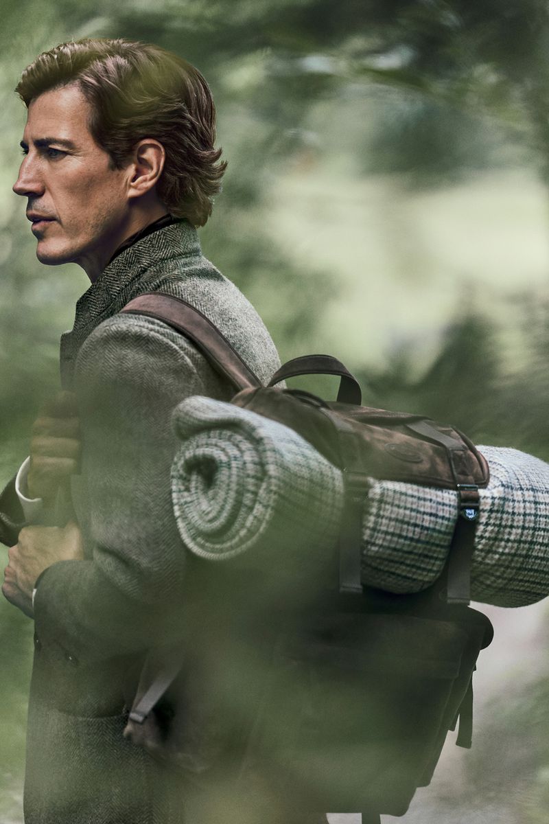 Adult male in a tweed coat carries a backpack in a wooded area, with blurred foliage in the foreground and filtered sunlight.