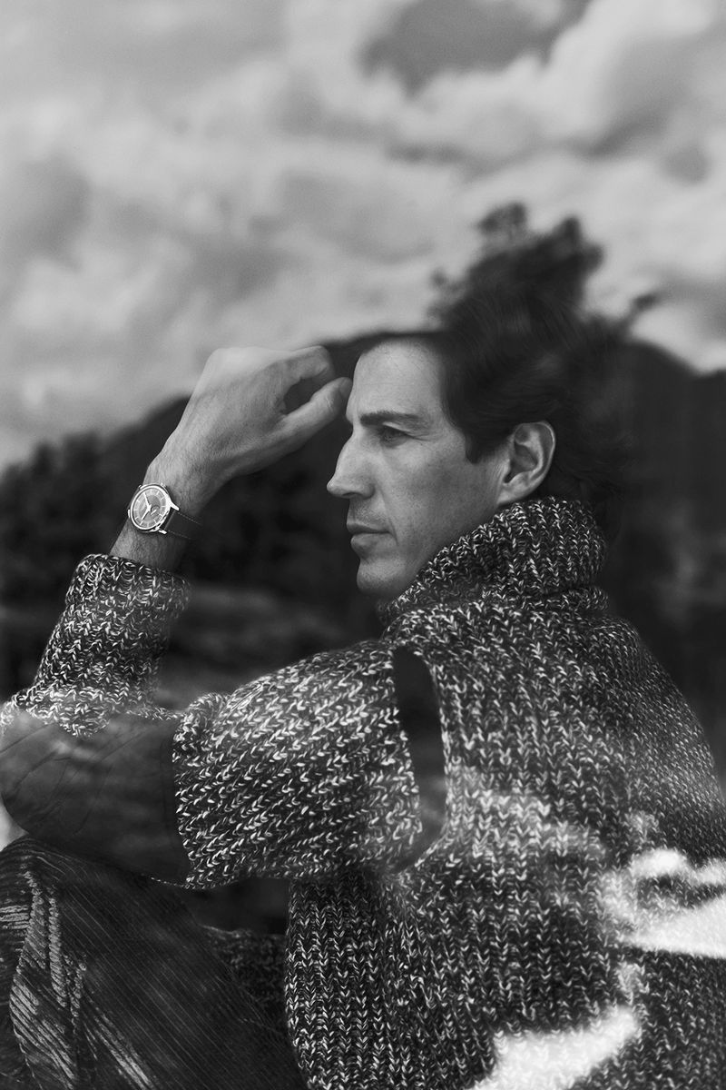Monochrome portrait of adult man in knitted sweater with mountains and clouds in the background, wearing wristwatch and hand on head.