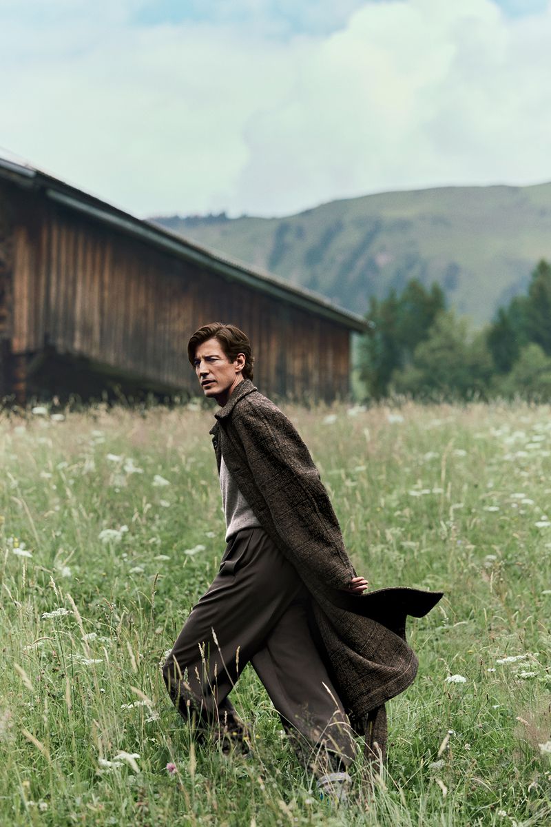 A slender man in an overcoat and trousers poses in a grassy field near a wooden building with mountains in the background on an overcast day.