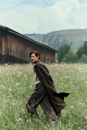 A slender man in an overcoat and trousers poses in a grassy field near a wooden building with mountains in the background on an overcast day.