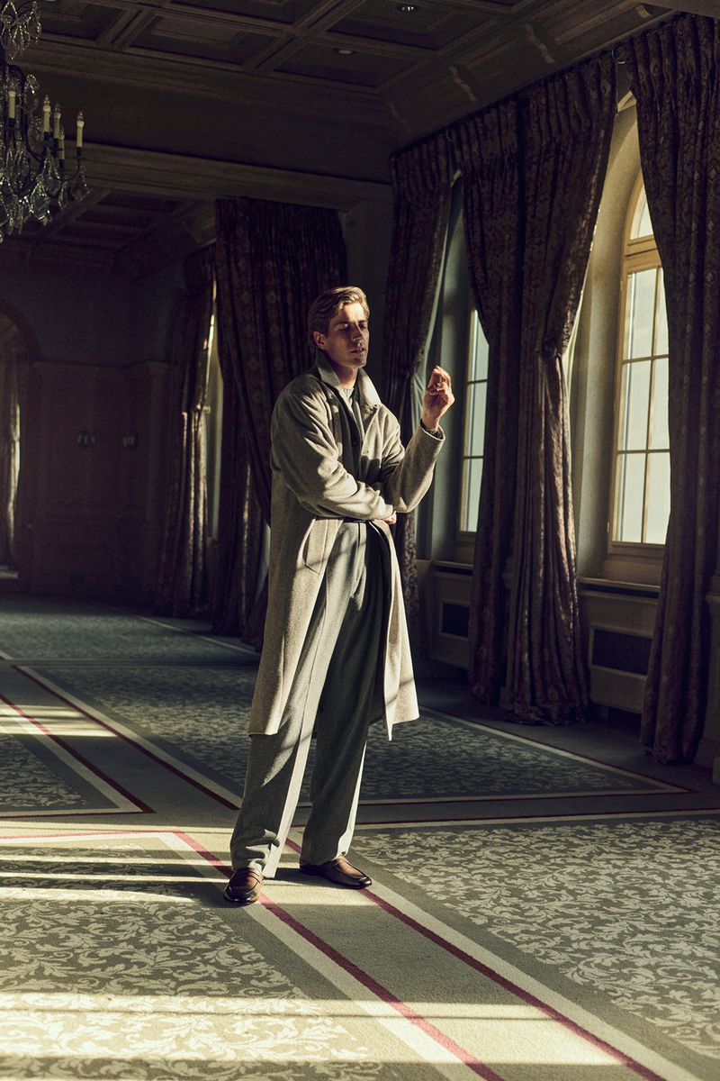 A man in a trench coat standing in a grand hall with large windows and patterned floors, bathed in soft, diffused natural light.