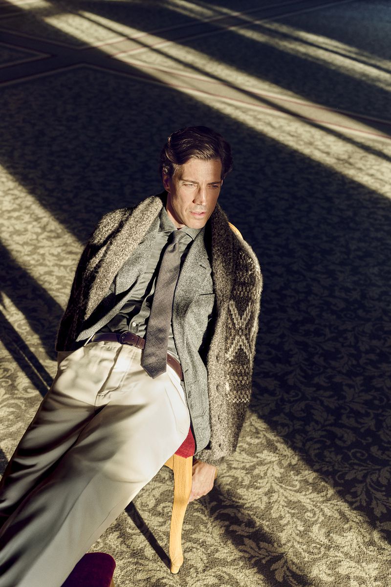 Man sitting in sunlight on a patterned rug wearing a cardigan sweater, dress shirt, tie, and dress pants.