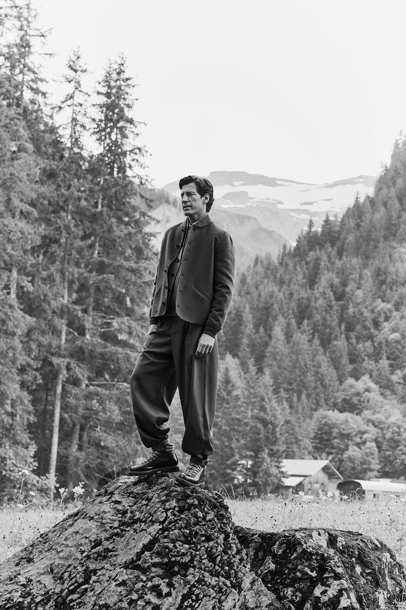 Adult male poses on a rock formation wearing vintage-style suit outdoors with background of pine trees and distant snow-capped mountains.