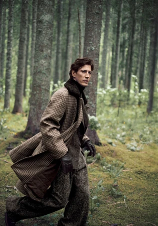 Model Oriol Elcacho walks through a mossy pine forest wearing a brown houndstooth overcoat and textured tweed trousers.
