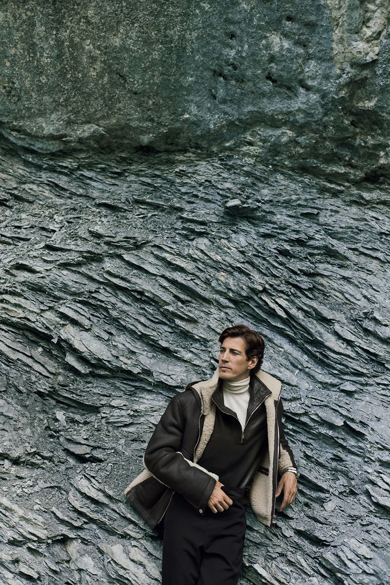 Man in shearling jacket leans against a rock formation, looking off camera in a medium shot. Fashion editorial outdoors.