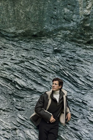 Man in shearling jacket leans against a rock formation, looking off camera in a medium shot. Fashion editorial outdoors.