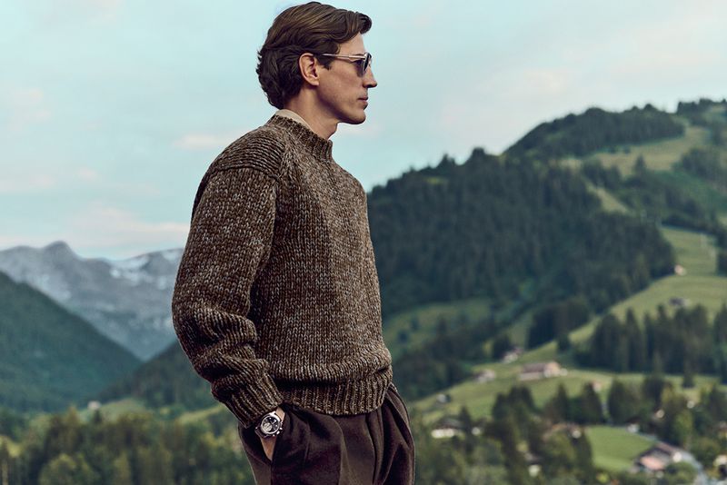 Adult male wearing a brown knit sweater, sunglasses and watch, stands in front of a green mountain landscape in a medium shot.