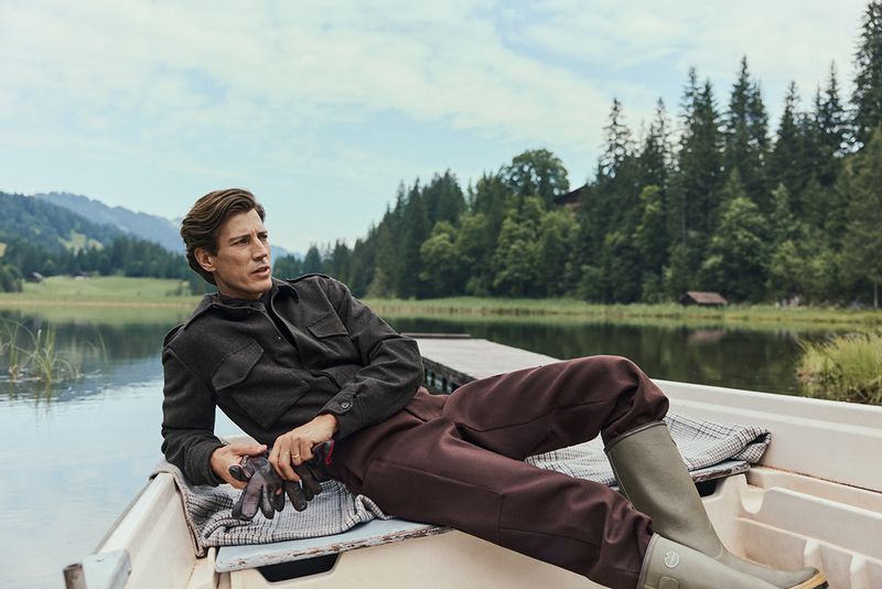 Man in boat on lake surrounded by forest. He wears a dark jacket, brown pants, and boots, holding gloves while relaxing.