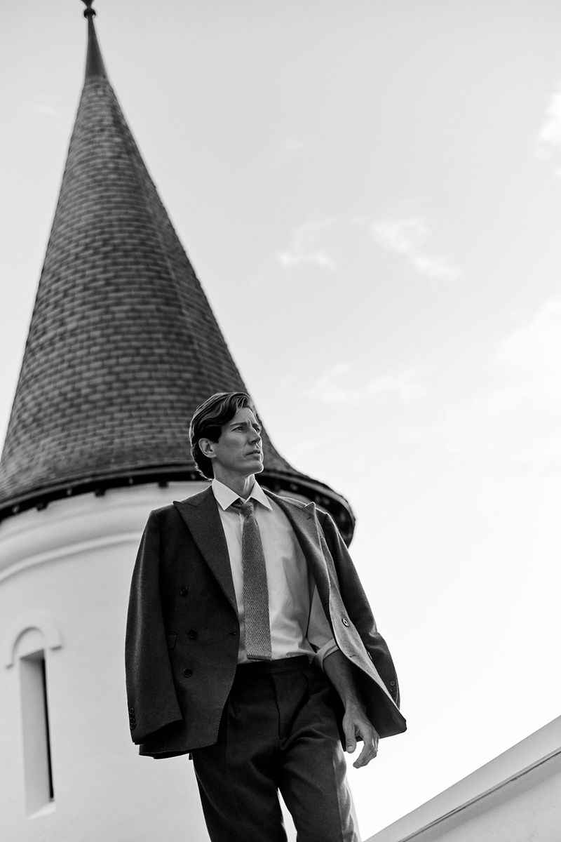 Monochrome portrait of an adult man in a suit with a building featuring a conical tower set against a bright sky background.