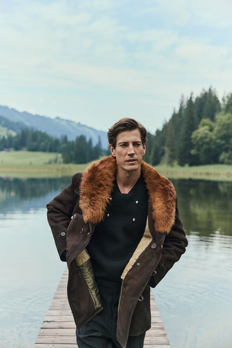 Male model in a fur-collared coat stands on a wooden dock by a serene lake surrounded by mountains and trees in the daytime.